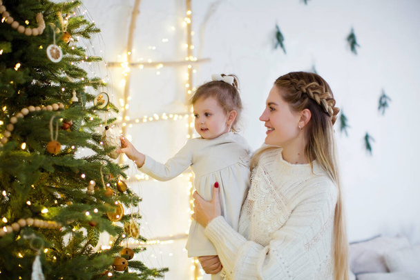 Young mother and daughter near Christmas tree. Happy family waiting for Christmas and New year. Cozy light interior with Christmas decorations and lights - Zdjęcie, obraz
