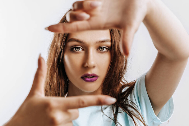 Beautiful young girl makes a frame with her hands on an isolated gray background. The woman with fashion make-up with purple lips seriously looking at the camera. Place for advertising. - Фото, зображення