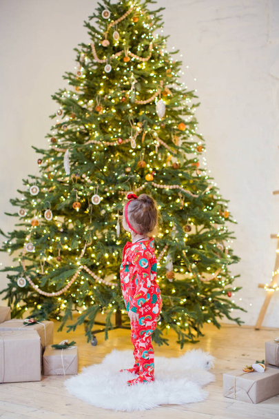 Aranyos kislány piros pizsamában áll közel karácsonyfa díszek és fények - Fotó, kép