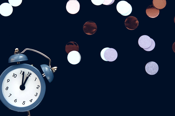 El reloj despertador vintage azul muestra a las 12 en punto aislado sobre fondo oscuro con un brillante metafán redondo de confeti. Despierta y date prisa. Cuenta atrás para la medianoche del año nuevo. - Foto, imagen