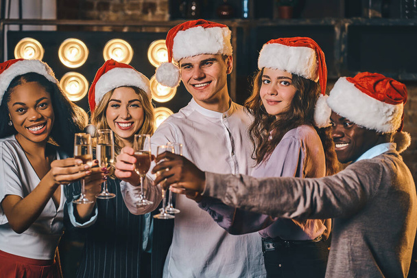 Smiling friends wearing Santa hats while holding glasses of champagne and clinking. Happy New Year concept - Zdjęcie, obraz
