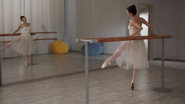 Υπέροχη χορεύτρια στην αίθουσα μπαλέτου κάνει κινήσεις με τα πόδια της στο πλάι και μπροστά, στέκεται μπροστά στον καθρέφτη - Φωτογραφία, εικόνα