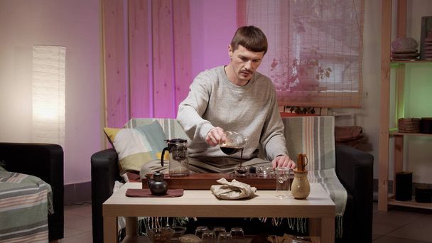 Στο σπίτι, ένας νεαρός χύνει τσάι από μια κανάτα σε ένα ποτήρι και πίνει φρέσκο τσάι. - Φωτογραφία, εικόνα