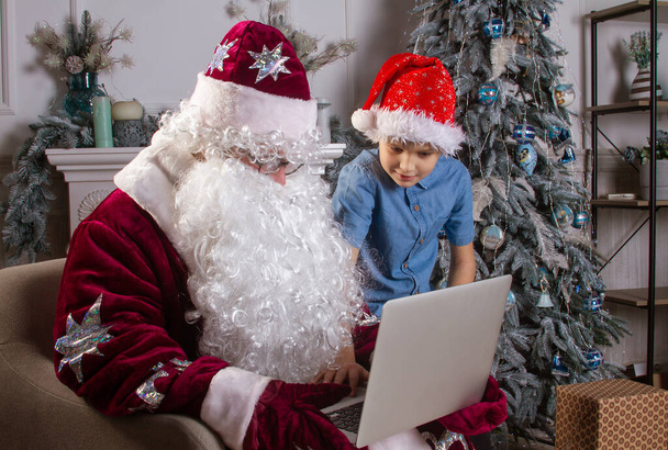 Santa Claus and a boy in a red hat are sitting by the Christmas tree, the child looks into Santa's bag and takes out gifts, Christmas and New Year, winter holidays. - Foto, Imagem