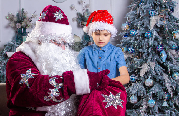 Santa Claus and a boy in a red hat are sitting by the Christmas tree, the child looks into Santa's bag and takes out gifts, Christmas and New Year, winter holidays. - Fotoğraf, Görsel
