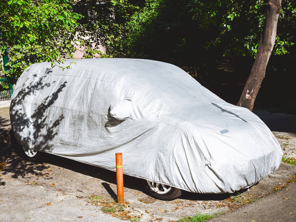 L'auto coperta da una copertura nel cortile sotto gli alberi illuminati dalla luce del sole - Foto, immagini