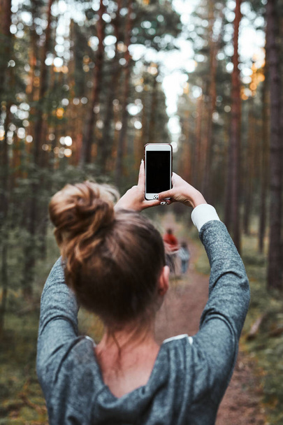Young woman taking photos using smartphone during walk in a forest on summer vacation trip. Back view of girl holding smartphone - Фото, изображение