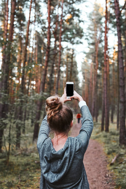 Young woman taking photos using smartphone during walk in a forest on summer vacation trip. Back view of girl holding smartphone - Foto, afbeelding