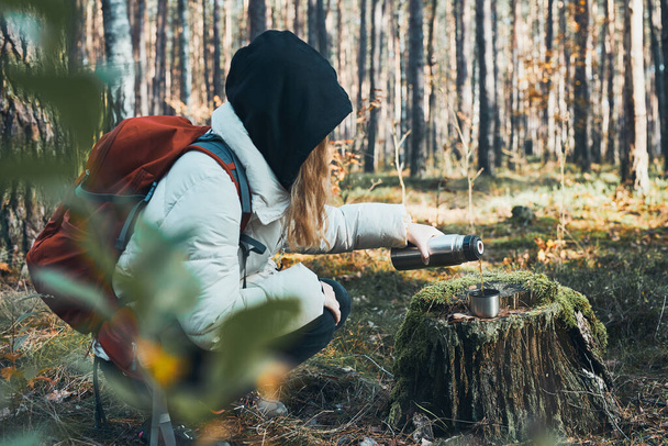 Woman having break during autumn trip pouring a hot drink from thermos flask on autumn cold day. Active women wandering in a forest actively spending time - Foto, afbeelding