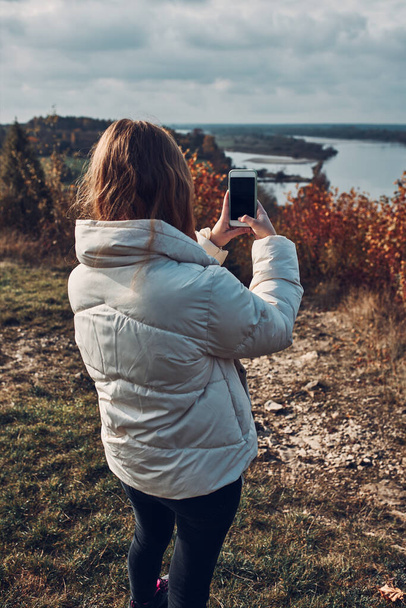 Back view of young woman taking photos of landscape with smartphone during trip on autumn sunny day - Foto, afbeelding