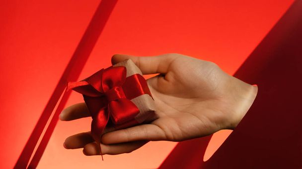 Lidská ruka drží dárkovou krabici zdobenou červenou stuhou a řemeslným papírem přes papír broušený kruh na červeném pozadí. Vánoční svátky, Silvestr. Dárková poukázka. Nápis - prodej, zavřít - Fotografie, Obrázek