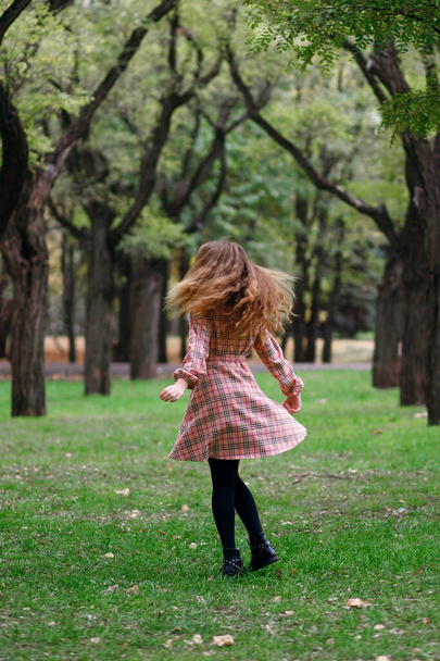An emotional girl with red hair runs along a green lawn in the park. The child is dressed in a checkered pink dress and black shoes. The girl rejoices and waves her hands cheerfully. Vertical photo - Photo, Image