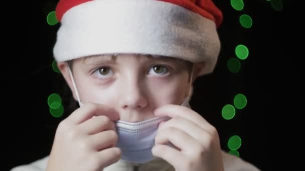 petite fille en rouge Santa Claus chapeau met sur masque de protection médicale, gants bleus - Séquence, vidéo