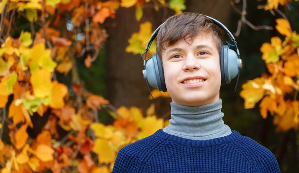 portrét teenagera poslouchajícího hudbu sluchátky, relaxace v podzimním městském parku, jasně žluté javorové listy jako pozadí - Fotografie, Obrázek