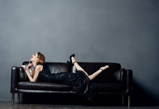 vonzó nő fekete ruhában fekszik egy bőr kanapén beltérben emelt lábakkal - Fotó, kép