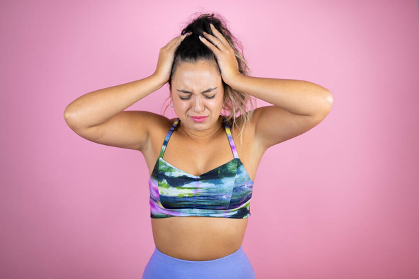 Jonge mooie vrouw draagt sportkleding over geïsoleerde roze achtergrond lijdt aan hoofdpijn wanhopig en gestrest omdat pijn en migraine met haar handen op het hoofd - Foto, afbeelding