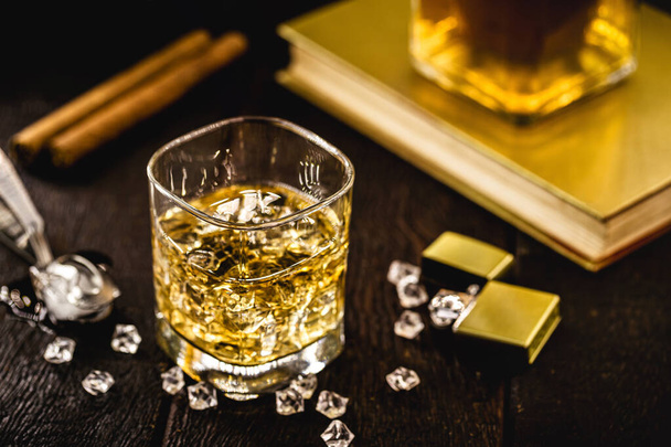 whisky com gelo em uma mesa de madeira, com objetos decorativos no fundo, charuto, isqueiro e livro. - Foto, Imagem
