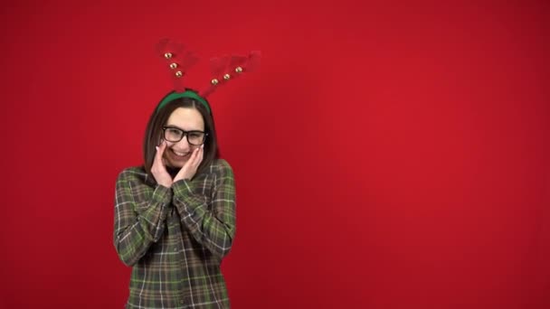 Een jonge vrouw staat met een hoofdband in de vorm van kersthoorns en is verlegen. Studio fotografie op een rode achtergrond. - Video