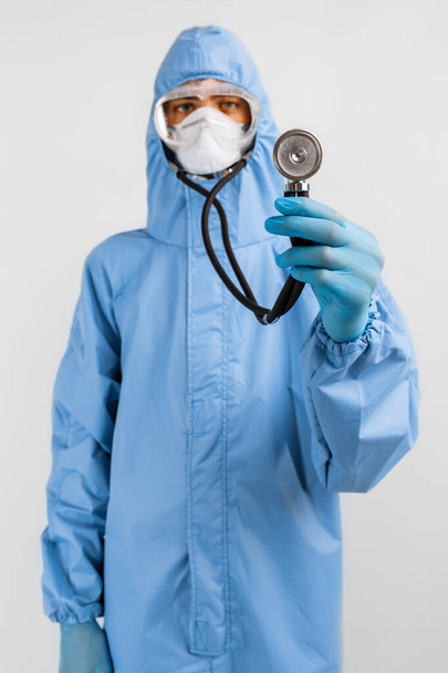 Doctor in a protective suit against coronavirus, mask, glasses and gloves is holding a stethoscope, listening to something, Medicine and healthcare concept - Photo, Image