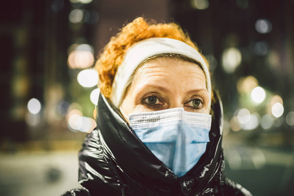Volwassen vrouw met gezichtsmasker bescherming, oudere vrouw in gezichtsmasker als gevolg van luchtvervuiling in de avondstad als achtergrond, zieke oude mensen met medisch masker. Verontreiniging, stofallergie en gezondheidsconcept - Foto, afbeelding