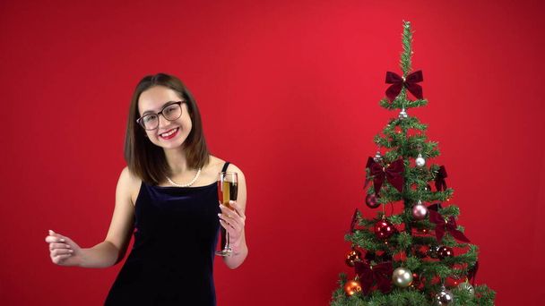 Jeune femme dansant avec une coupe de champagne près du sapin de Noël sur fond rouge. Une fille avec des lunettes et une robe de soirée. - Photo, image