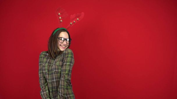 Mladá žena stojí s čelenkou v podobě vánočních rohů a stydí se. Studio fotografie na červeném pozadí. - Fotografie, Obrázek