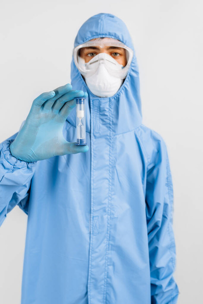 doctor in a protective suit and a medical mask on his face, looking at an hourglass with sand flowing down, limited time, coronavirus - Foto, afbeelding