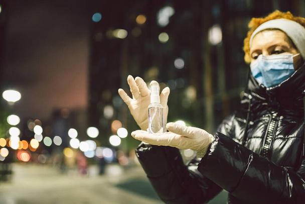 保護コロナウイルスとcovid 19.冬の夜の街の路上で成熟した女性の手の感染症に対する防腐剤。マスクと蜂蜜手袋の高齢女性は病気の収縮を恐れている. - 写真・画像