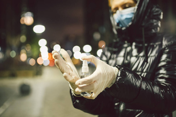 防腐、衛生、ヘルスケアの概念。新しい正常よ。手に消毒剤を持っている成熟した白人女性,保護医療マスクや手袋を身に着けている夜の都市の背景に立って. - 写真・画像