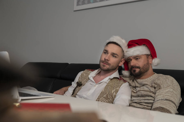 Joven pareja gay caucásica con sombreros de Santa Claus viendo películas en el portátil en la víspera de Navidad - Foto, Imagen