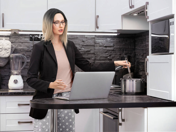 Ξανθιά κοπέλα με γυαλιά, μαύρο επίσημο σακάκι και παντελόνι πιτζάμας διεξάγει επιχειρηματικές συνομιλίες στο Διαδίκτυο μέσω ενός φορητού υπολογιστή στην κουζίνα και ανάδευση σε μια κατσαρόλα στο φούρνο, λειτουργεί εξ αποστάσεως στο σπίτι - Φωτογραφία, εικόνα