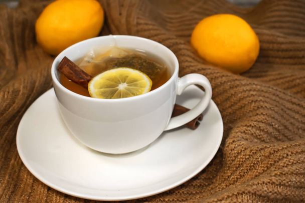 Detailní záběr bílého poháru s čajovým citrónem a skořicí, který stojí na teplém žlutém svetru na pozadí rozmazaných citronů. - Fotografie, Obrázek