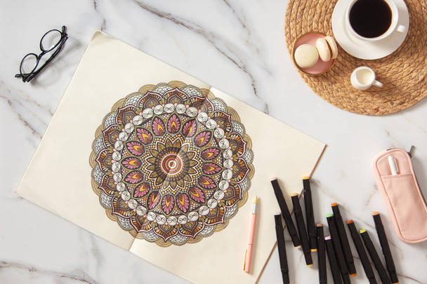 Woman drawing and coloring mandala in sketchbook with colorful markers while having breakfast with coffee and macarons on white marble table. Stress relieving trend by painting. Art therapy. Leisure. - Photo, image