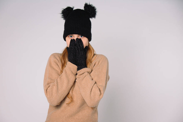 A blue-eyed girl in black winter hat and gloves holds her hands to her face. Grey background with space. High quality photo - Photo, Image