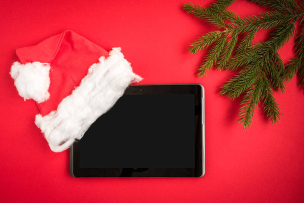 Οθόνη tablet με χριστουγεννιάτικο καπέλο και κλαδιά ελάτης σε κόκκινο φόντο. - Φωτογραφία, εικόνα