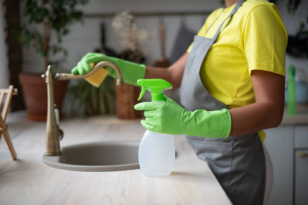 Algemene schoonmaak van de keuken. Een vrouw in een schort en rubberen handschoenen wast de gootsteen. Professionele huishoudelijke dienst. Veegt de kraan af met een doek - Foto, afbeelding