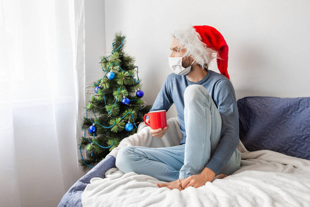 Bożonarodzeniowe popijawy Covid-19 i pandemia New Normal. Młody człowiek w masce na twarz i kapelusz Mikołaja siedzący na kanapie, trzymający czerwony kubek kawy, sam, przed choinką w salonie. - Zdjęcie, obraz