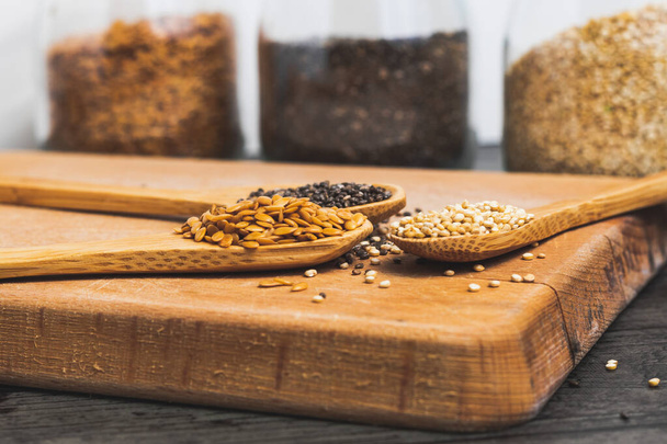 Drei Holzlöffel mit Flachs, Quinoa und Chia-Samen umrahmen einen braunen rustikalen Tisch und im Hintergrund stehen drei Glasbecher mit Flachs, Quinoa und Chia-Samen. - Foto, Bild