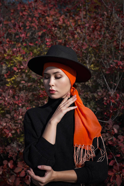 Gyönyörű fiatal nő sétál az őszi erdőben. Meleg napos idő. Őszi koncepció. divatos portré ázsiai nőről kalapban és narancssárga sállal a fején. - Fotó, kép