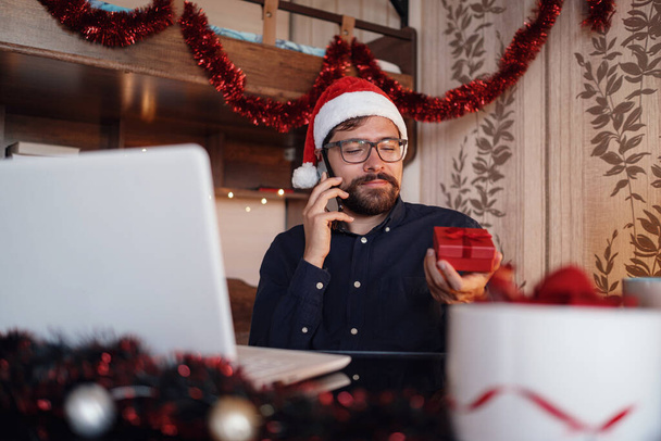 Közelkép portré vidám kaukázusi jóképű férfi santa kalap ül a hangulatos szobában Xmas dekoráció hívja okostelefon boldog arcát. Újév. Boldog karácsonyt! - Fotó, kép