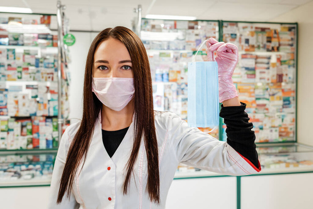 A female pharmacist in a medical mask and gloves, holding a medical mask in her hand. In the background, the pharmacy Windows are blurred. Concept of protection against coronavirus. - Photo, Image