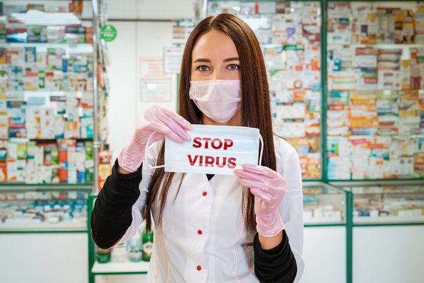 Zatrzymać wirus.Kobieta farmaceuta w masce medycznej i rękawiczki, trzymając maskę medyczną w rękach. W tle okna apteki są zamazane. Pojęcie ochrony przed wirusami i chorobami. - Zdjęcie, obraz