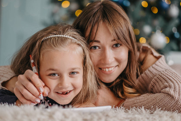 Mom helps her daughter write a letter to Santa near the Christmas tree at home. - 写真・画像