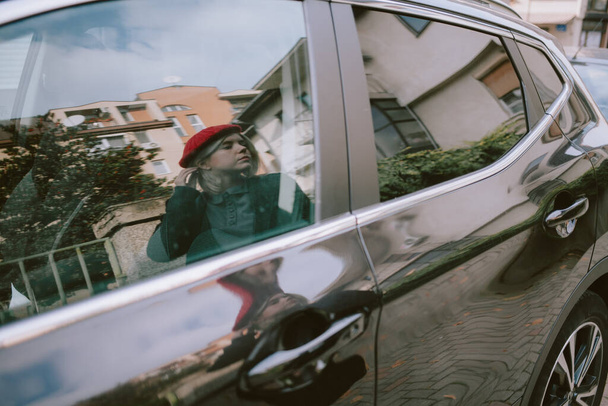 Молодая женщина с красным беретом идет по улице и проверяет свое отражение в окне машины - Фото, изображение