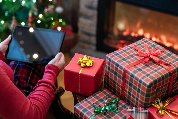 Christmas. Woman in sweater using tablet for searching gift ideas sitting at table near fireplace and christmas tree. Concept - Foto, Imagen
