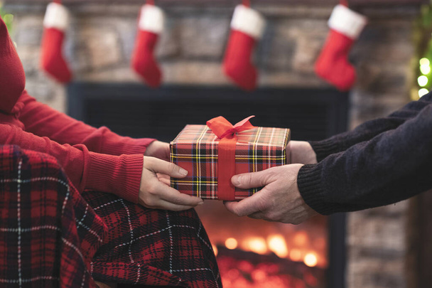 Man giving Christmas and New Year Gift box to woman in front of the fireplace with flame.  - 写真・画像