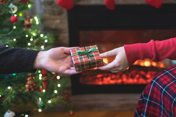 Man giving Christmas and New Year Gift box to woman in front of christmas tree, fireplace with flame. Xmas concept. - Фото, изображение