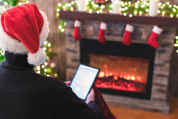 Christmas. Man in santa claus hat with tablet sitting near christmas tree and fireplace. Concept of online service in holidays. Back view. - Photo, Image