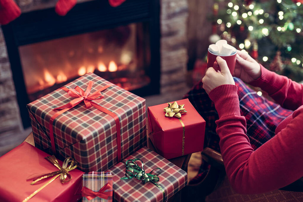 Woman with red mug of hot chocolate and melted marshmallow packing handmade christmas gifts sitting near christmas tree with and fireplace. - Фото, зображення