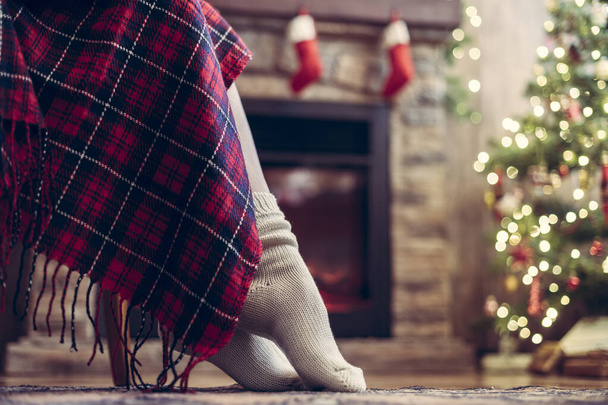 Bottom view of woman legs in knitted winter socks sitting on armchair covered plaid near christmas tree and fireplace. Bottom view  - Foto, Imagem